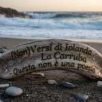 Un pezzo di legno levigato con la scritta incisa "Non Versi di Iolanda La Carruba - Questa non è una poesia" poggia sulla sabbia di una spiaggia con il mare sullo sfondo.